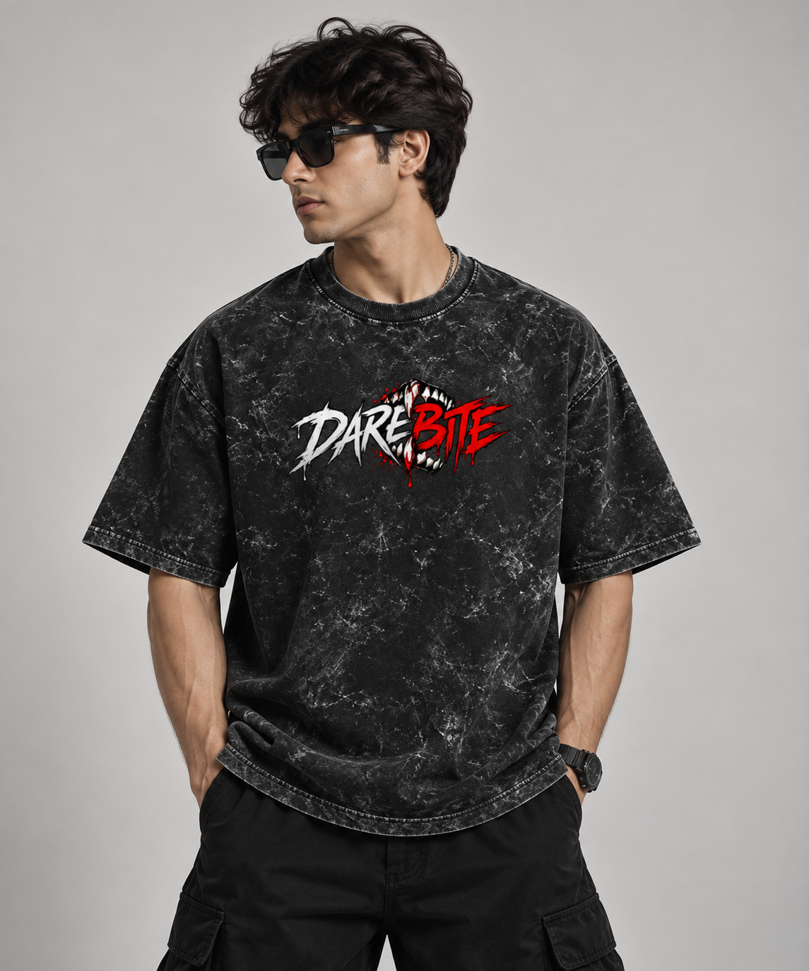 A man wearing a black acid-wash graphic t-shirt with a 'DARE BITE' logo on the chest, paired with black cargo pants and sunglasses, against a solid gray studio background.