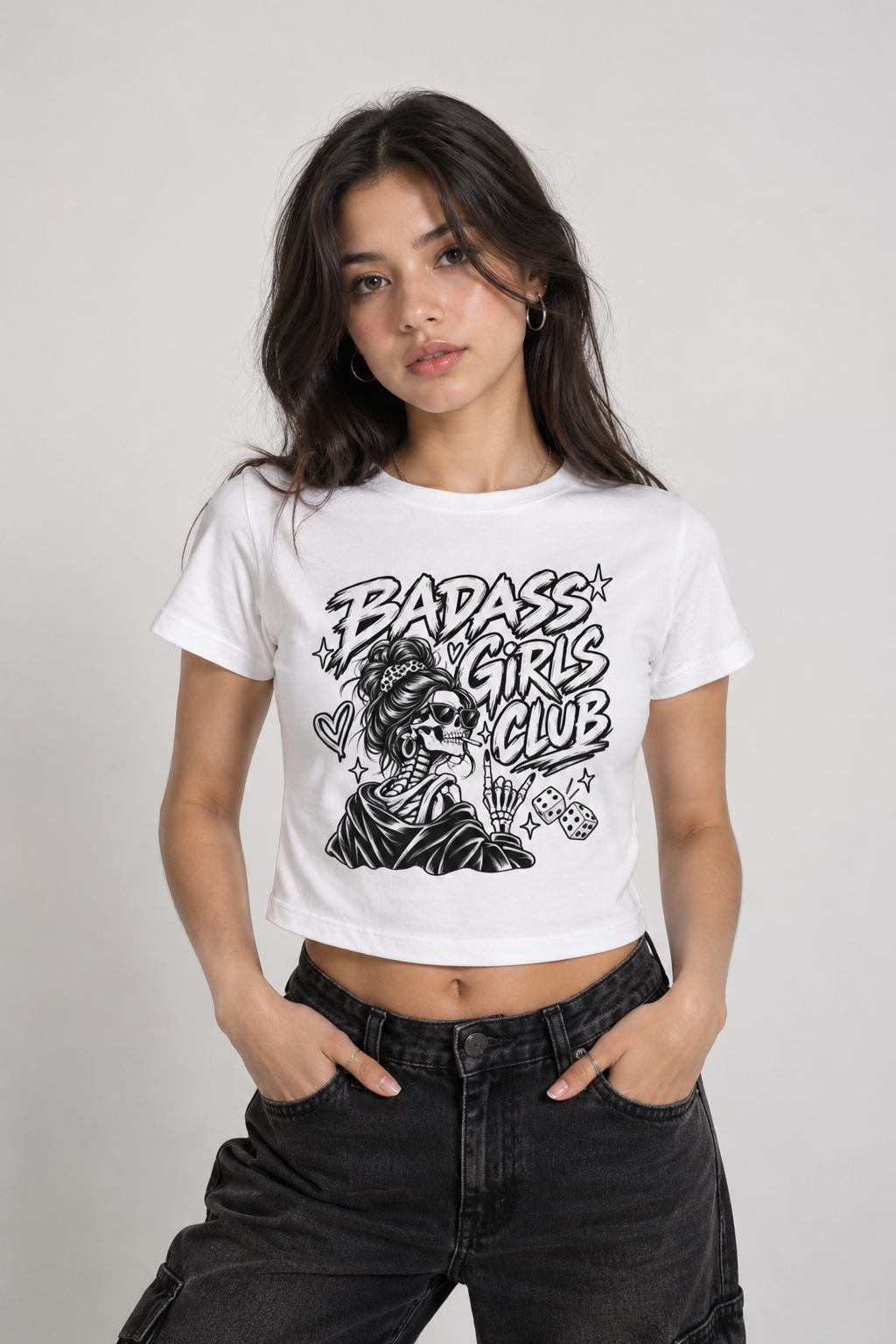 A woman modeling a white cropped graphic t-shirt with a black 'BADASS GIRLS CLUB' skull illustration, paired with black jeans, against a light gray studio background.