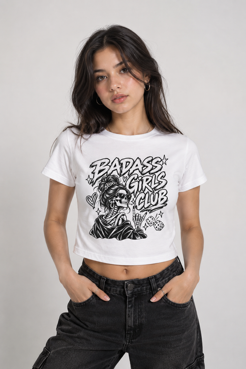 A woman modeling a white cropped graphic t-shirt with a black 'BADASS GIRLS CLUB' skull illustration, paired with black jeans, against a light gray studio background.