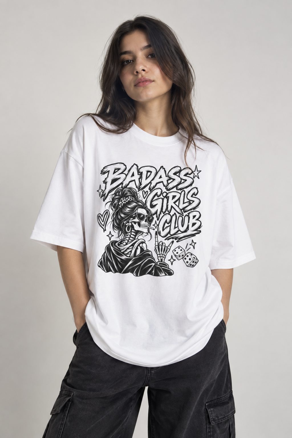 A woman wearing a white oversized graphic t-shirt with a black 'Badass Girls Club' skeleton illustration, paired with black cargo pants, against a light gray studio background.