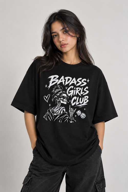 A woman wearing a black oversized graphic t-shirt with a white 'BADASS GIRLS CLUB' text and skeleton illustration, paired with dark cargo pants, against a light gray studio background.