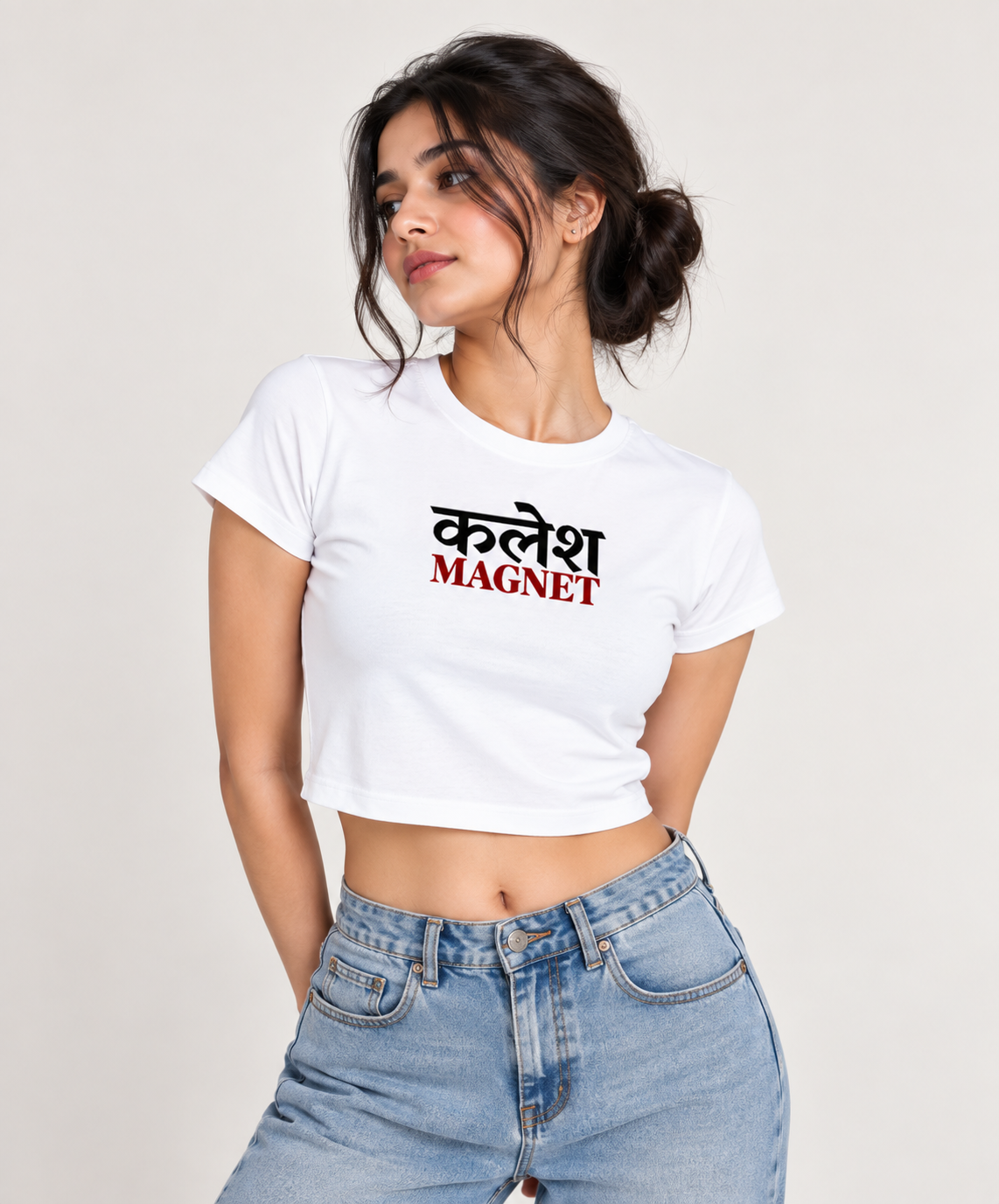 A woman wearing a white crop top t-shirt with the Hindi text 'कलेश MAGNET' printed on the chest, paired with light blue jeans, against a plain light background.