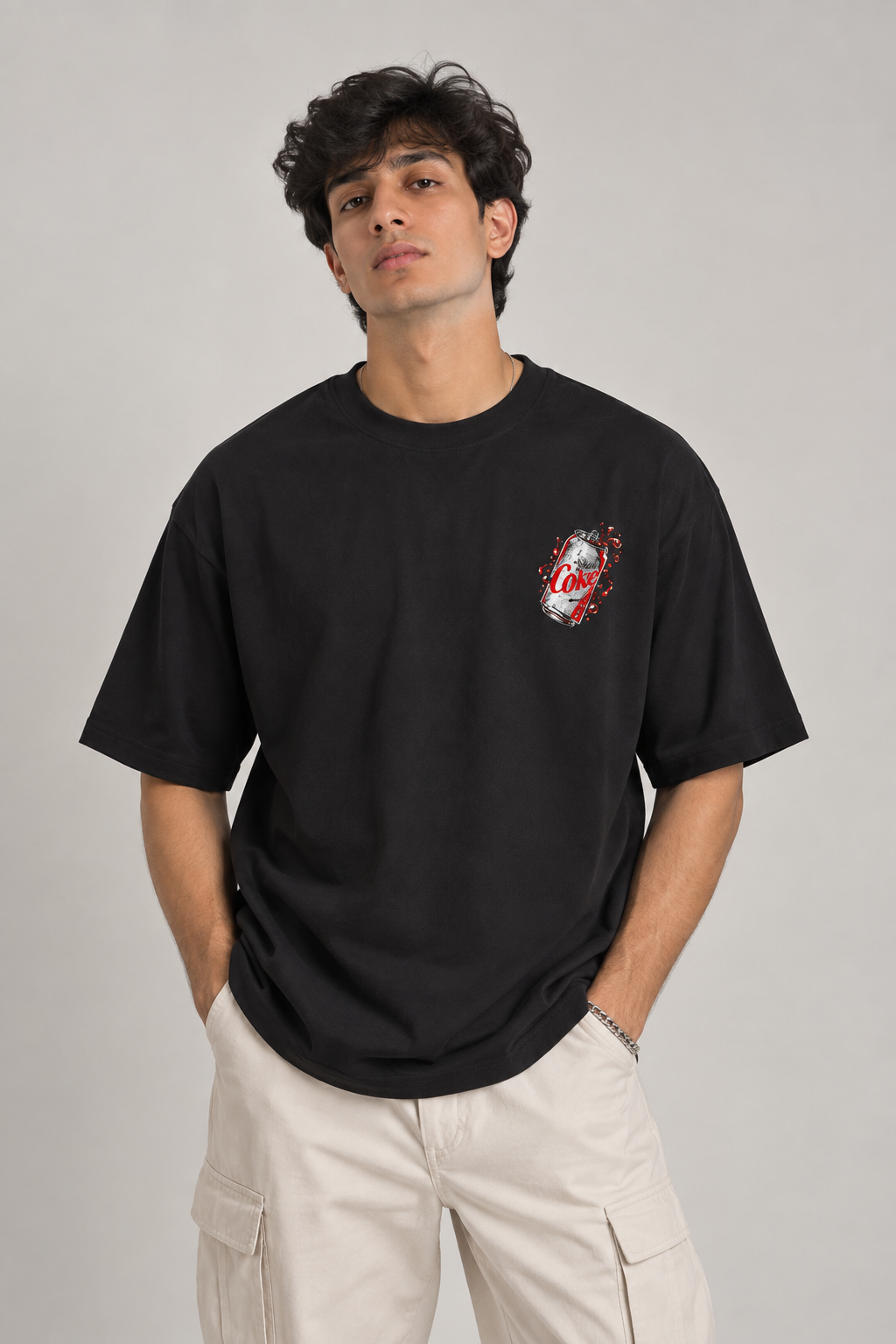 A man wearing a black short-sleeve t-shirt with a small red, white, and blue graphic on the left chest, paired with light beige cargo pants, against a solid gray studio background.