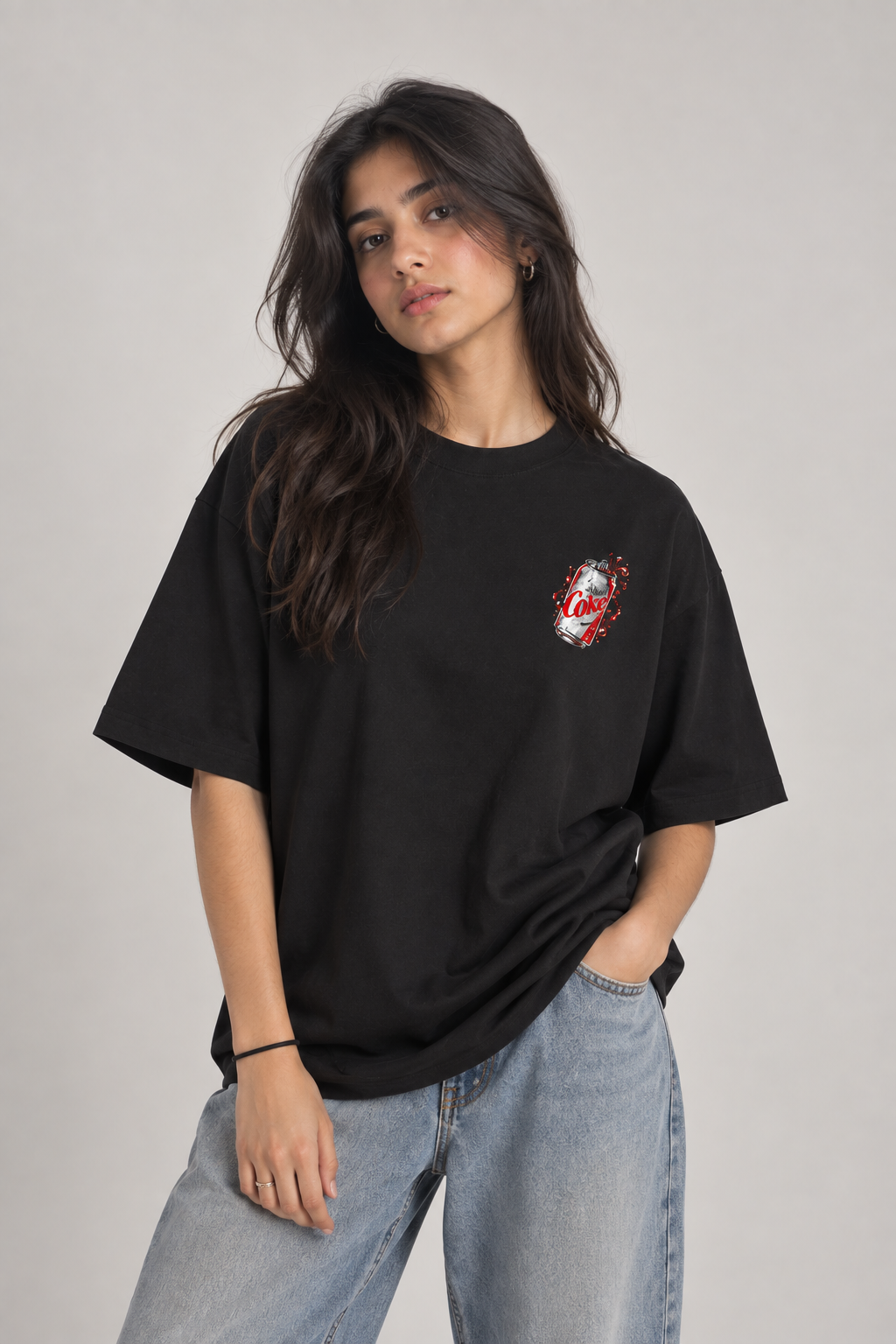 A woman wearing a black oversized t-shirt with a small red, white, and black graphic on the left chest, paired with light blue jeans, photographed against a neutral gray studio background.