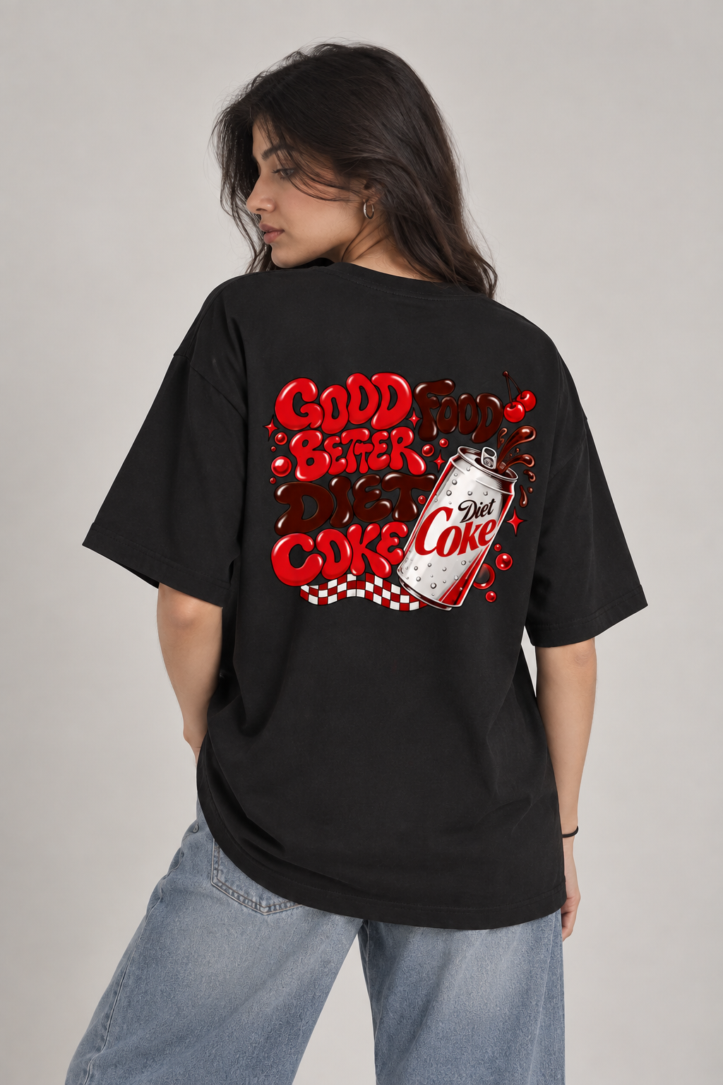 A woman wearing a black oversized t-shirt with a red and white retro 'Good Beer Bad Coke' graphic on the back, paired with light blue jeans, photographed against a neutral gray studio background.