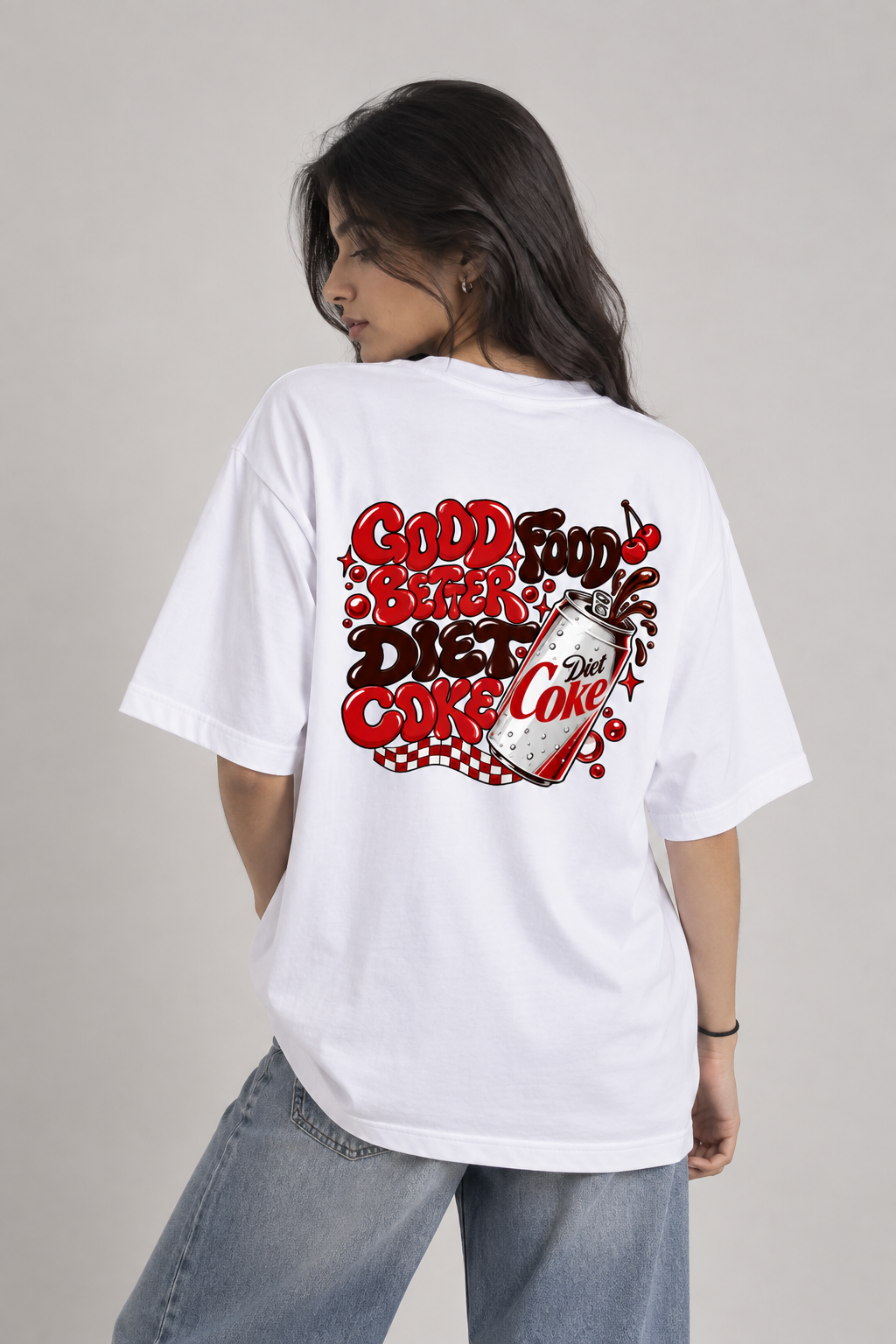 A woman wearing an oversized white graphic t-shirt with a red and black retro-style 'Good Food Diet Coke' print on the back, paired with light blue jeans, photographed against a neutral light gray background.