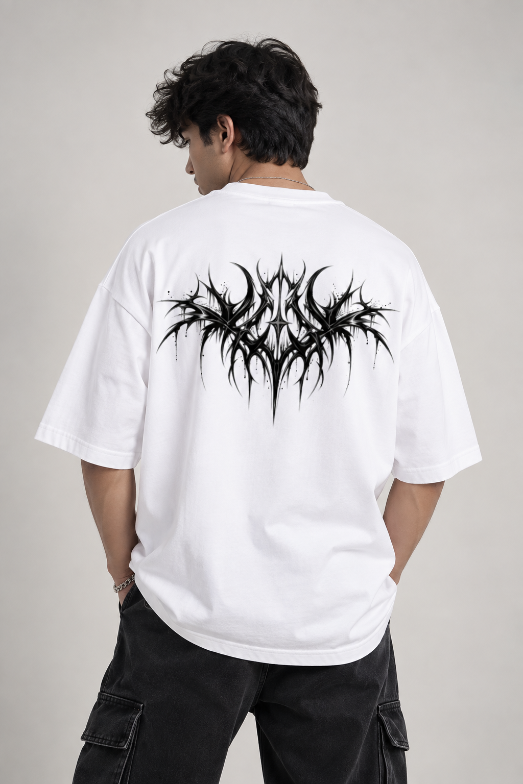 A man viewed from behind wearing a white oversized t-shirt with a large black spiky graphic on the back, paired with black cargo pants, against a plain light gray studio background.