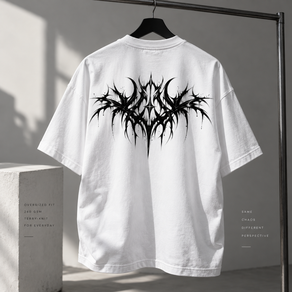 White oversized short-sleeve T-shirt hanging on a black hanger, shown from the back with a large black spiky graphic print across the upper back. The shirt is photographed indoors against a gray wall with dramatic shadows, next to a concrete block and beside a clothing rack.