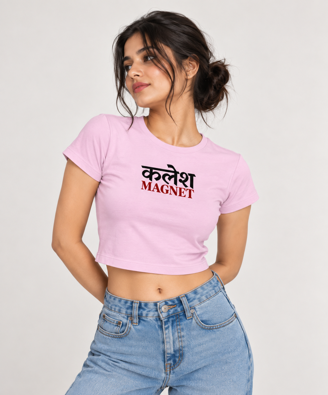 A woman wearing a pink crop top t-shirt with the Hindi text 'कलेश MAGNET' printed on the chest, paired with light blue jeans, against a plain light background.