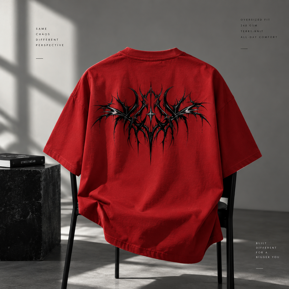 A red oversized short-sleeve T-shirt is draped over a black chair, showing the back with a large black spiky graphic print. The setting is a minimalist gray room with dramatic shadows, a dark block with a book, and decorative text on the sides.