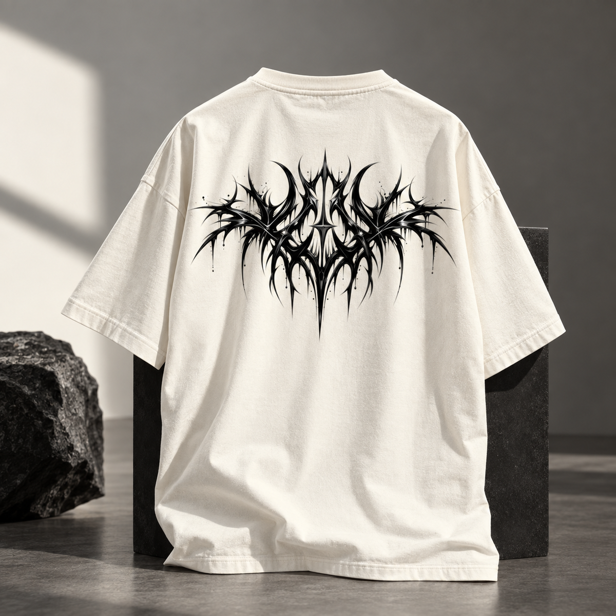 An off-white short-sleeve t-shirt shown from the back, featuring a large, detailed black spiky graphic print across the upper back. The shirt is displayed on a dark block in a styled studio setting with a rock prop on the left.