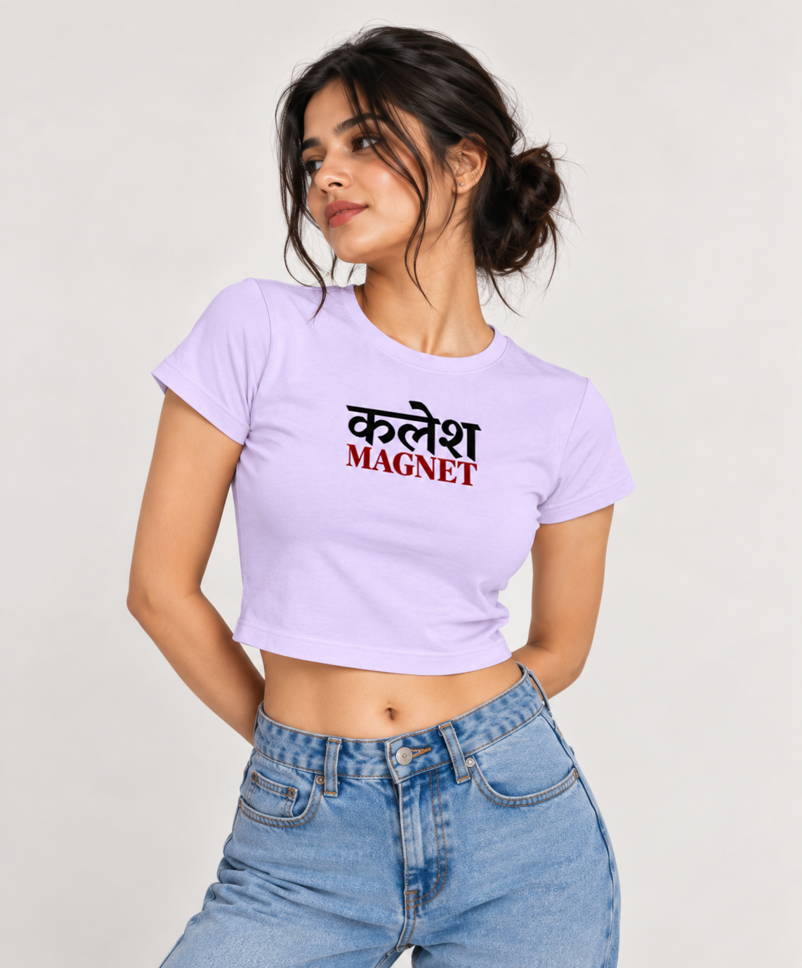 A woman wearing a lavendar crop top t-shirt with the Hindi text 'कलेश MAGNET' printed on the chest, paired with light blue jeans, against a plain light background.