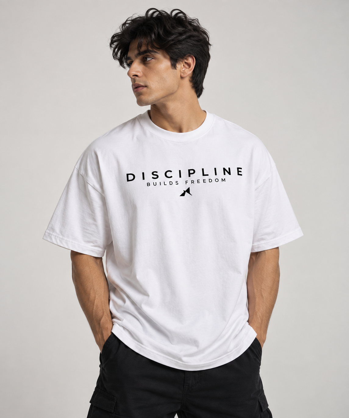 A man wearing a white short-sleeve t-shirt with a black graphic print that reads 'DISCIPLINE BUILDS FREEDOM' and a small silhouette of a person jumping, paired with black cargo pants, against a light gray studio background.