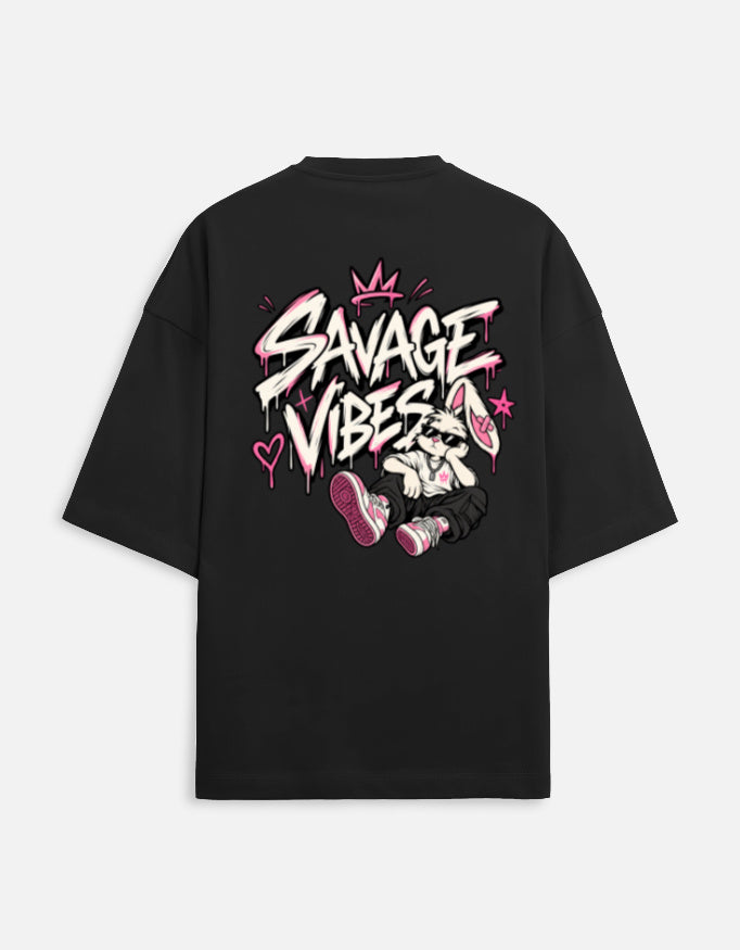 A black oversized short-sleeve t-shirt shown from the back, featuring a large graphic print with the words 'SAVAGE VIBES' in white and pink graffiti-style lettering, a pink crown, and a cartoon character wearing pink sneakers.