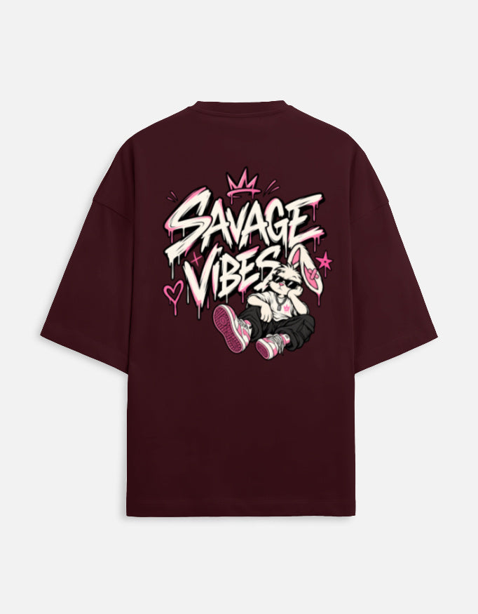 A dark maroon short-sleeve t-shirt shown from the back, featuring a large graphic print with the text 'SAVAGE VIBES' in white graffiti-style lettering, pink spray paint details, and a cartoon character sitting on the back.