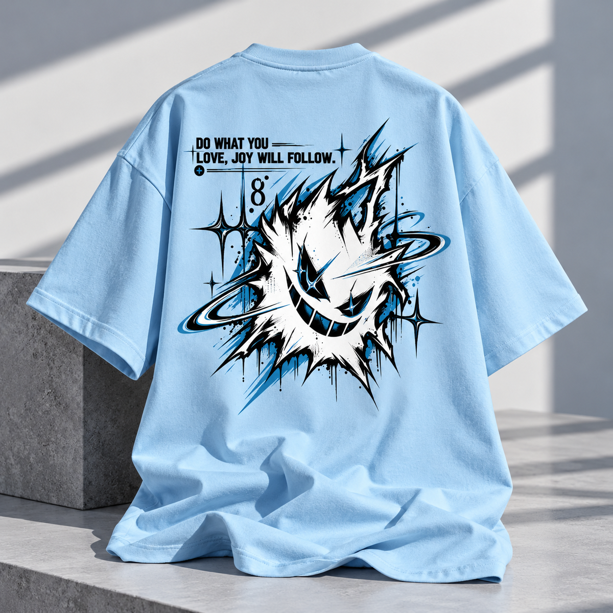 A light blue oversized T-shirt shown from the back, styled on a concrete ledge in a sunlit setting. The shirt features a large graphic print with a white and black abstract burst, blue accents, and the text 'DO WHAT YOU LOVE, JOY WILL FOLLOW.