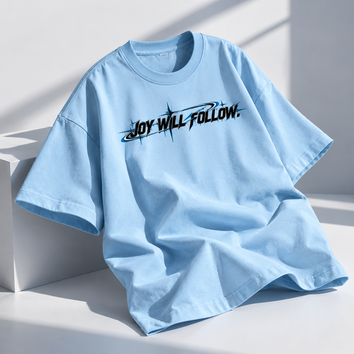 A light blue oversized short-sleeve t-shirt with a black graphic text print reading 'JOY WILL FOLLOW.' across the chest, styled on a white geometric platform with soft shadows.