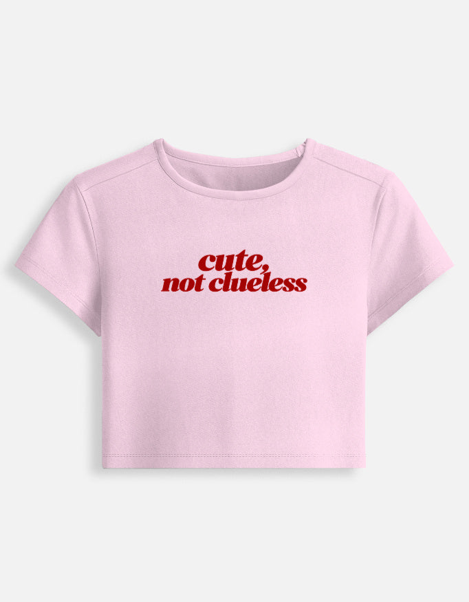 A light pink short-sleeve crop top t-shirt with the phrase 'cute, not clueless' printed in red across the chest, displayed on a clean off-white background.