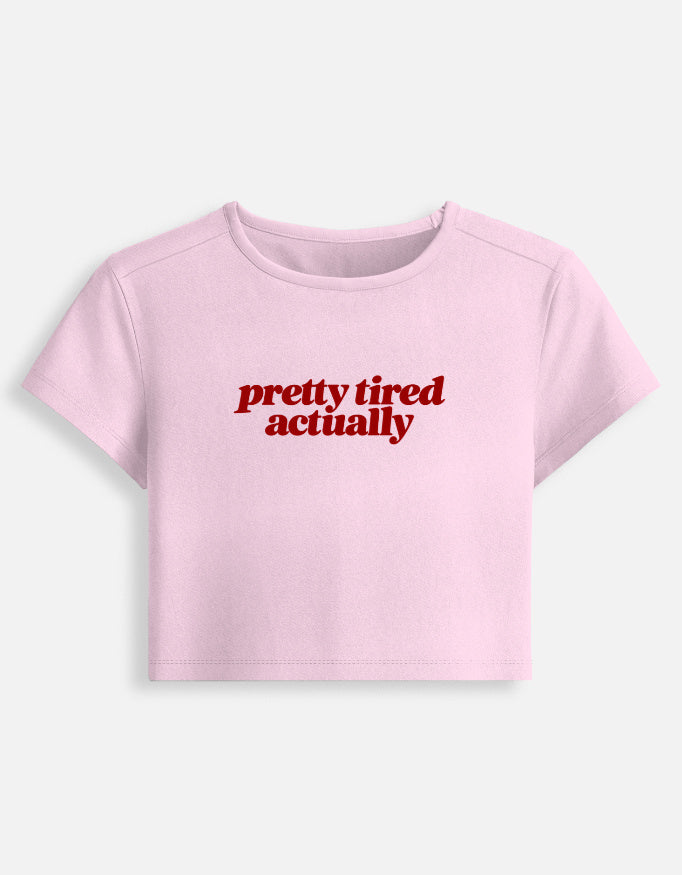 A light pink short-sleeve crop top t-shirt with the phrase 'pretty tired actually' printed in red serif text across the chest, displayed on a clean off-white background.