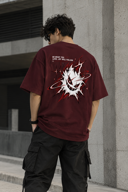 A man viewed from behind wearing a maroon t-shirt with a large white and red graphic print on the back, paired with black cargo pants, standing in front of a concrete building.