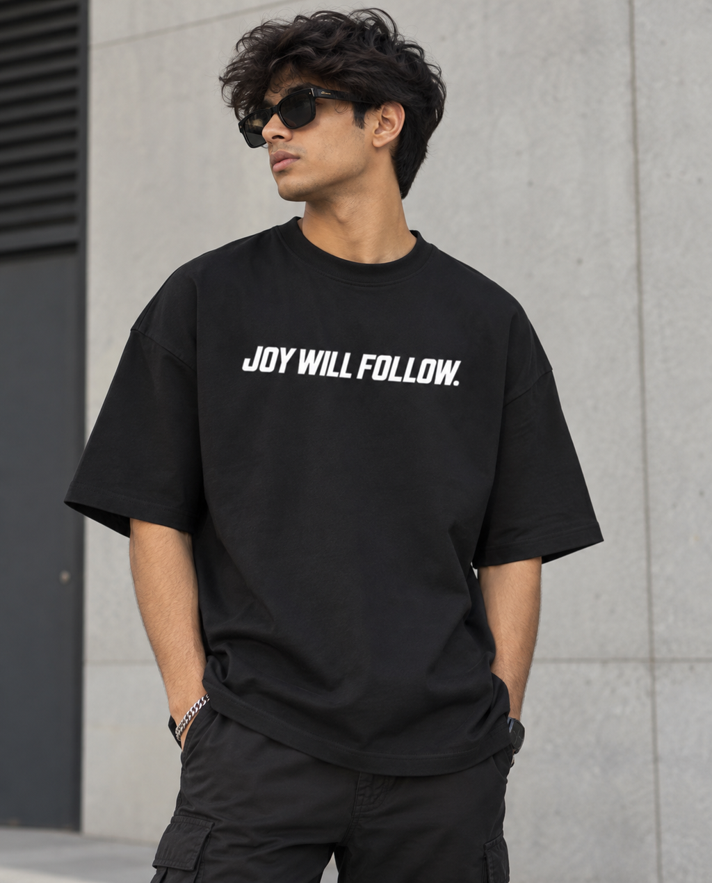 A man wearing a black oversized t-shirt with the white text 'JOY WILL FOLLOW.' across the chest, paired with black cargo pants and sunglasses, standing against a gray concrete wall.