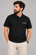 A man wearing a black short-sleeve polo shirt with 'No Rules' embroidered in gold on the left chest, paired with beige trousers, against a solid gray studio background.