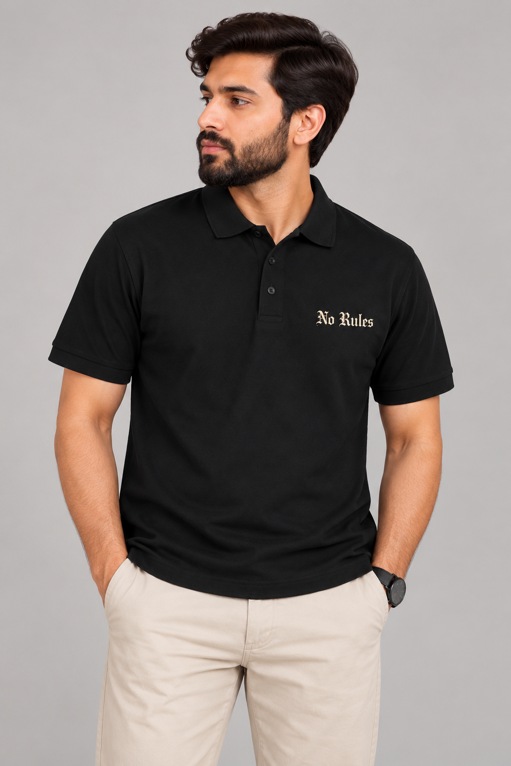 A man wearing a black short-sleeve polo shirt with 'No Rules' embroidered in gold on the left chest, paired with beige trousers, against a solid gray studio background.