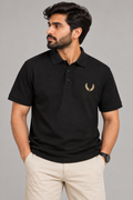 A man wearing a black short-sleeve polo shirt with a small gold embroidered laurel wreath logo on the left chest, paired with beige trousers, against a solid gray studio background.