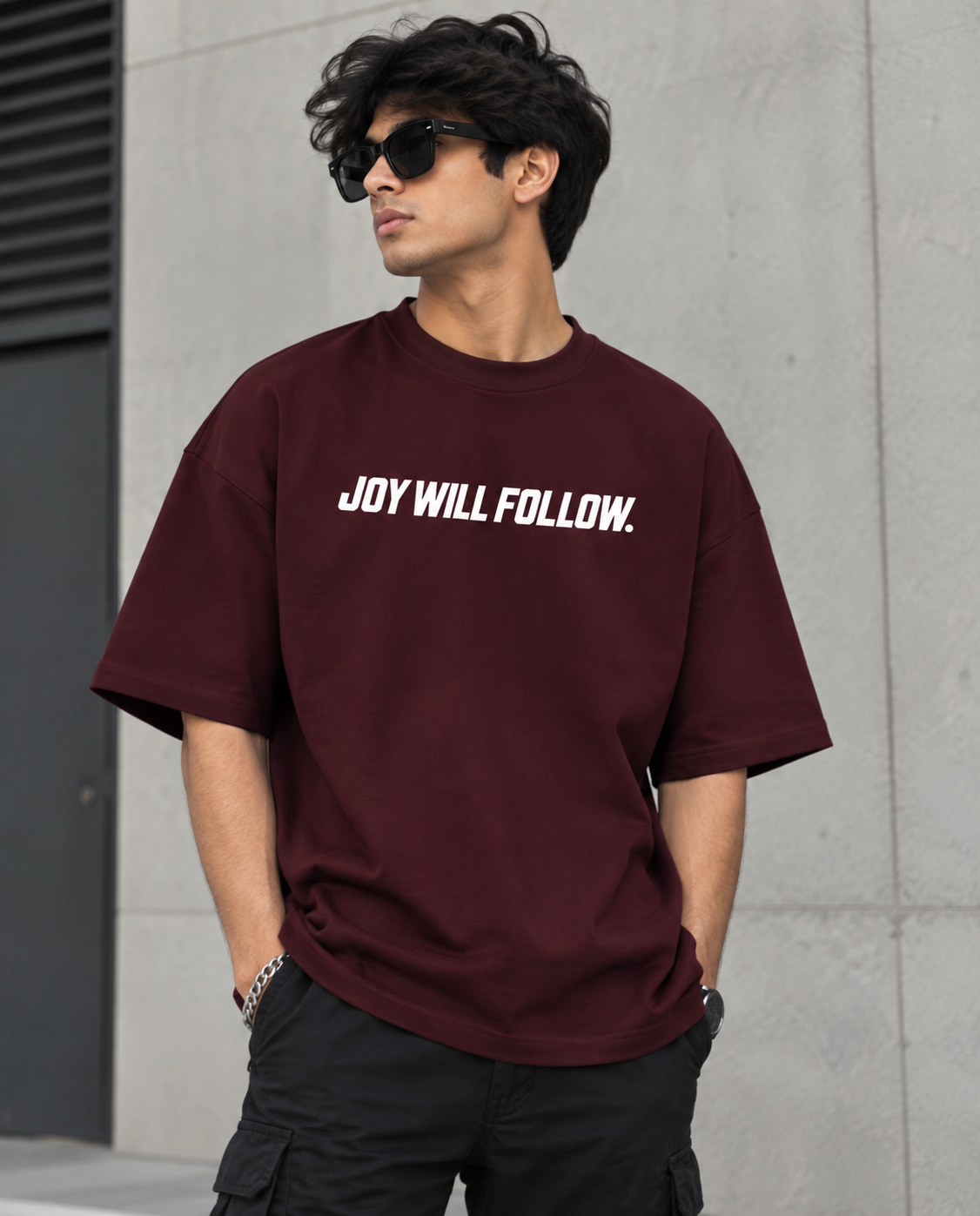 A man wearing a burgundy t-shirt with the white printed text 'JOY WILL FOLLOW.' on the chest, paired with black cargo pants and sunglasses, standing against a gray concrete wall.
