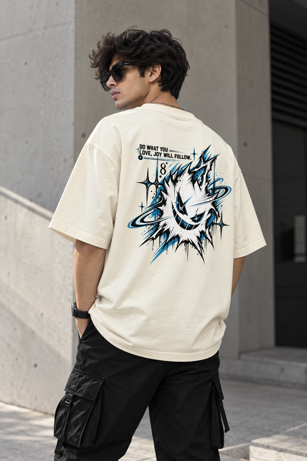 A man wearing a cream-colored oversized t-shirt with a large blue and black graphic print on the back, paired with black cargo pants, standing outdoors against a concrete wall.