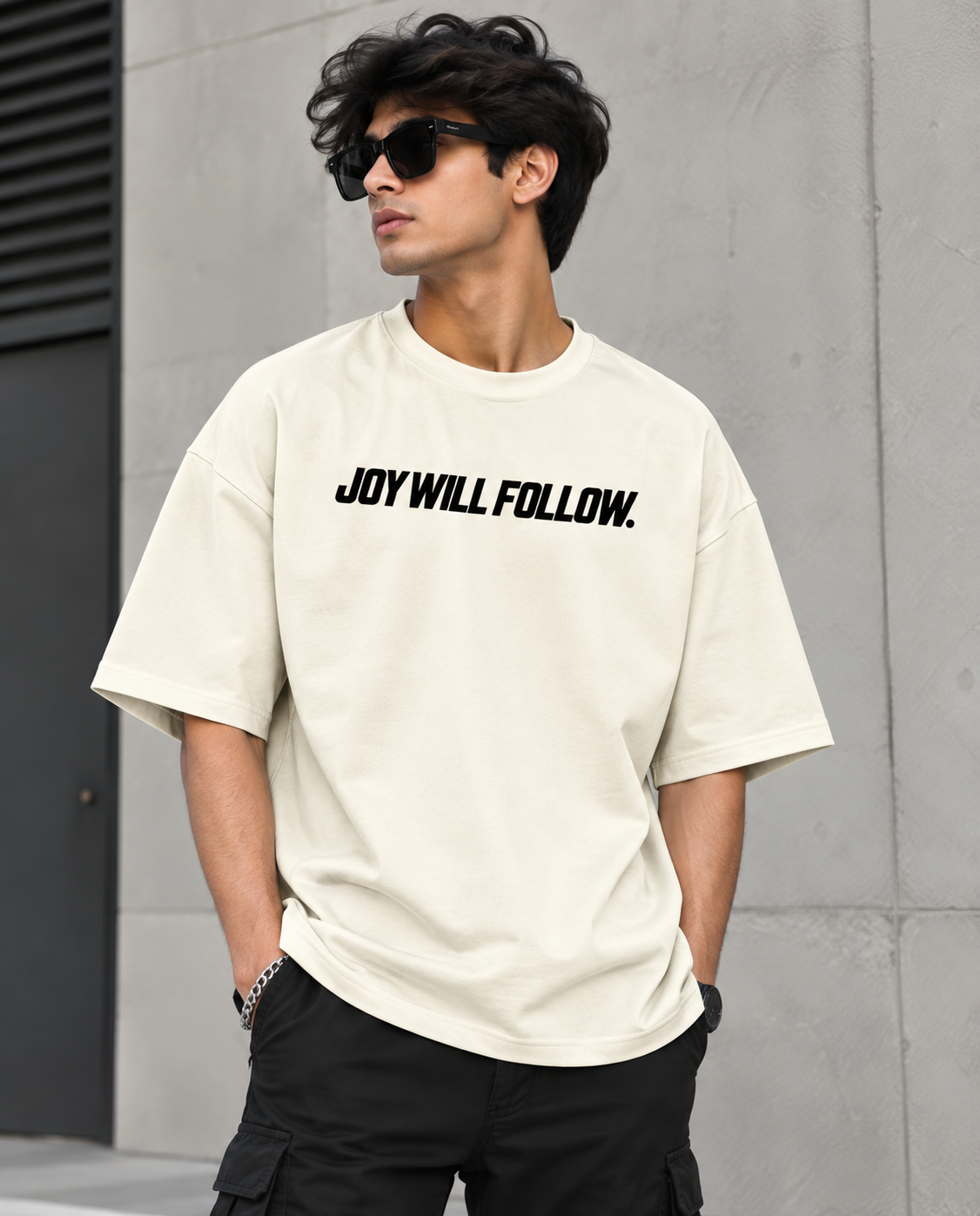 A man wearing a cream-colored oversized t-shirt with the black text 'JOY WILL FOLLOW.' printed across the chest, paired with black cargo pants and sunglasses, standing against a gray concrete wall.