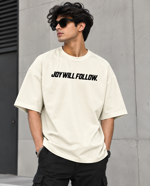 A man wearing a cream-colored oversized t-shirt with the black text 'JOY WILL FOLLOW.' printed across the chest, paired with black cargo pants and sunglasses, standing against a gray concrete wall.