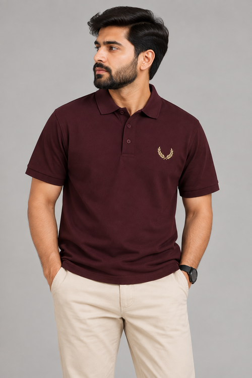 A man wearing a short-sleeve polo shirt in maroon with a small gold laurel leaf logo on the left chest, paired with beige trousers, against a solid gray studio background.