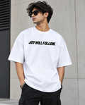 A man wearing a white crewneck t-shirt with the black text 'JOY WILL FOLLOW.' printed across the chest, paired with black cargo pants and sunglasses, standing against a gray concrete wall.