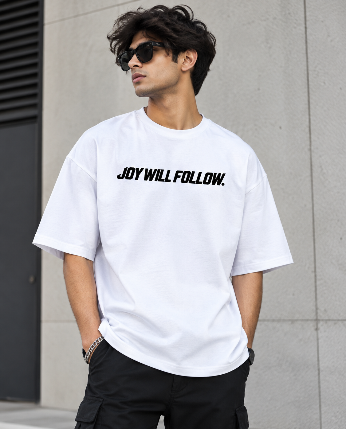 A man wearing a white crewneck t-shirt with the black text 'JOY WILL FOLLOW.' printed across the chest, paired with black cargo pants and sunglasses, standing against a gray concrete wall.
