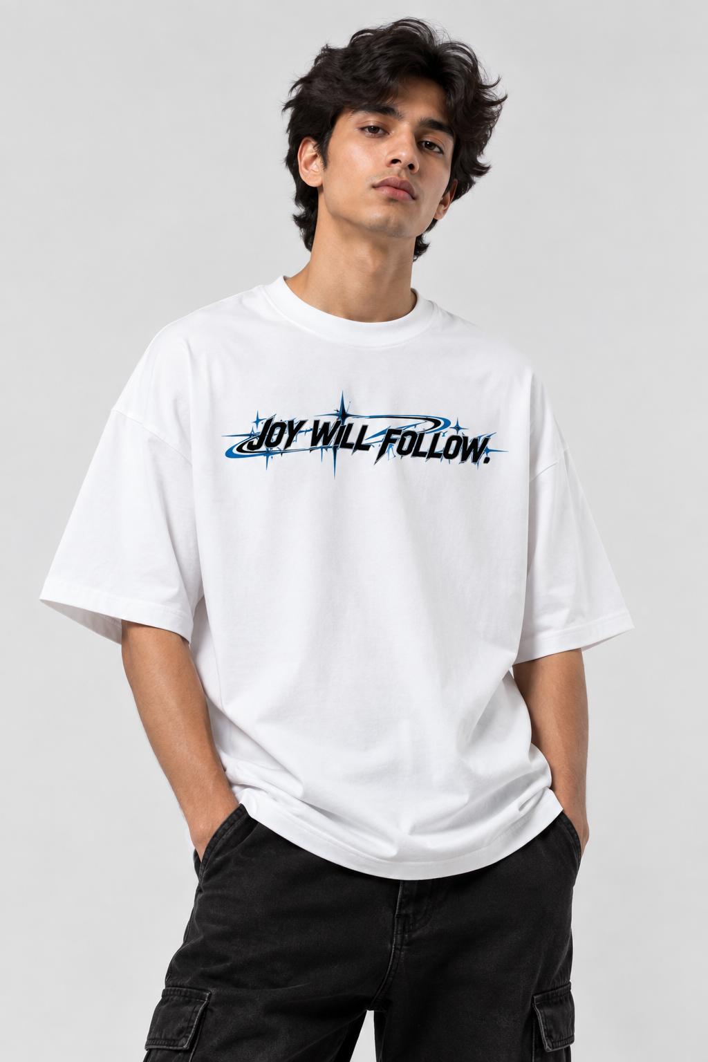 A man wearing a white oversized t-shirt with a black and blue graphic text 'JOY WILL FOLLOW.' on the chest, paired with black cargo pants, against a light gray studio background.