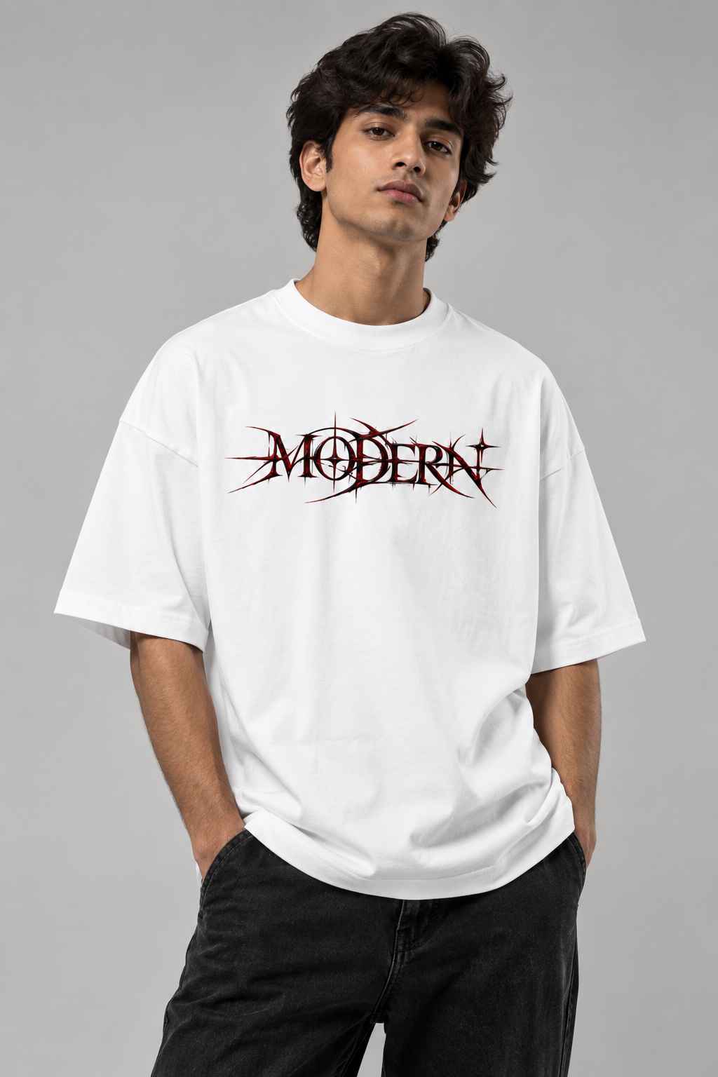 A man wearing a white oversized t-shirt with a stylized black and red 'MODERN' graphic across the chest, paired with black jeans, against a solid gray studio background.