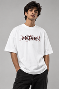 A man wearing a white oversized t-shirt with a stylized black and red 'MODERN' graphic across the chest, paired with black jeans, against a solid gray studio background.