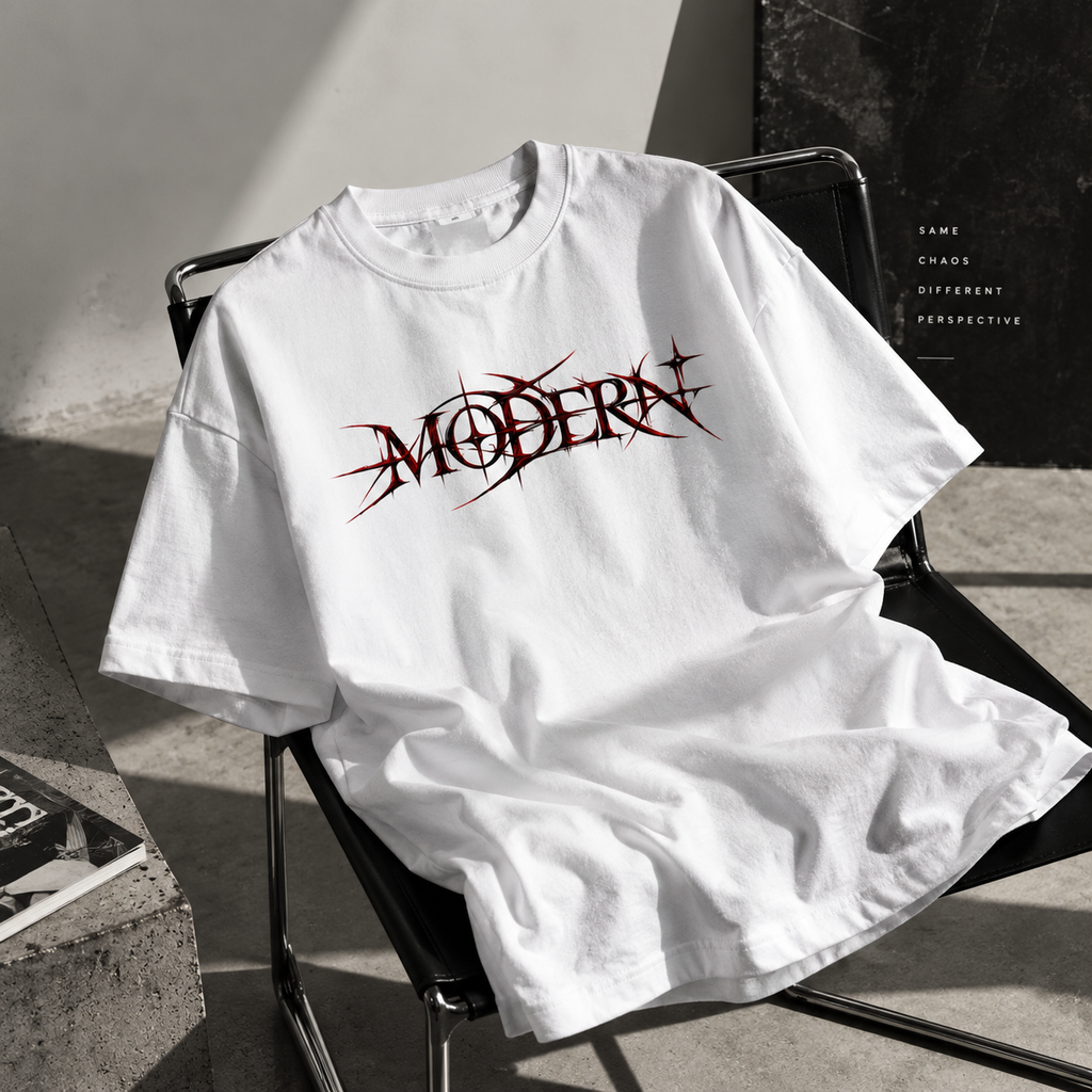 A white oversized T-shirt draped over a black metal chair in a sunlit studio setting. The shirt features a stylized dark red and black 'MODERN' graphic across the chest.