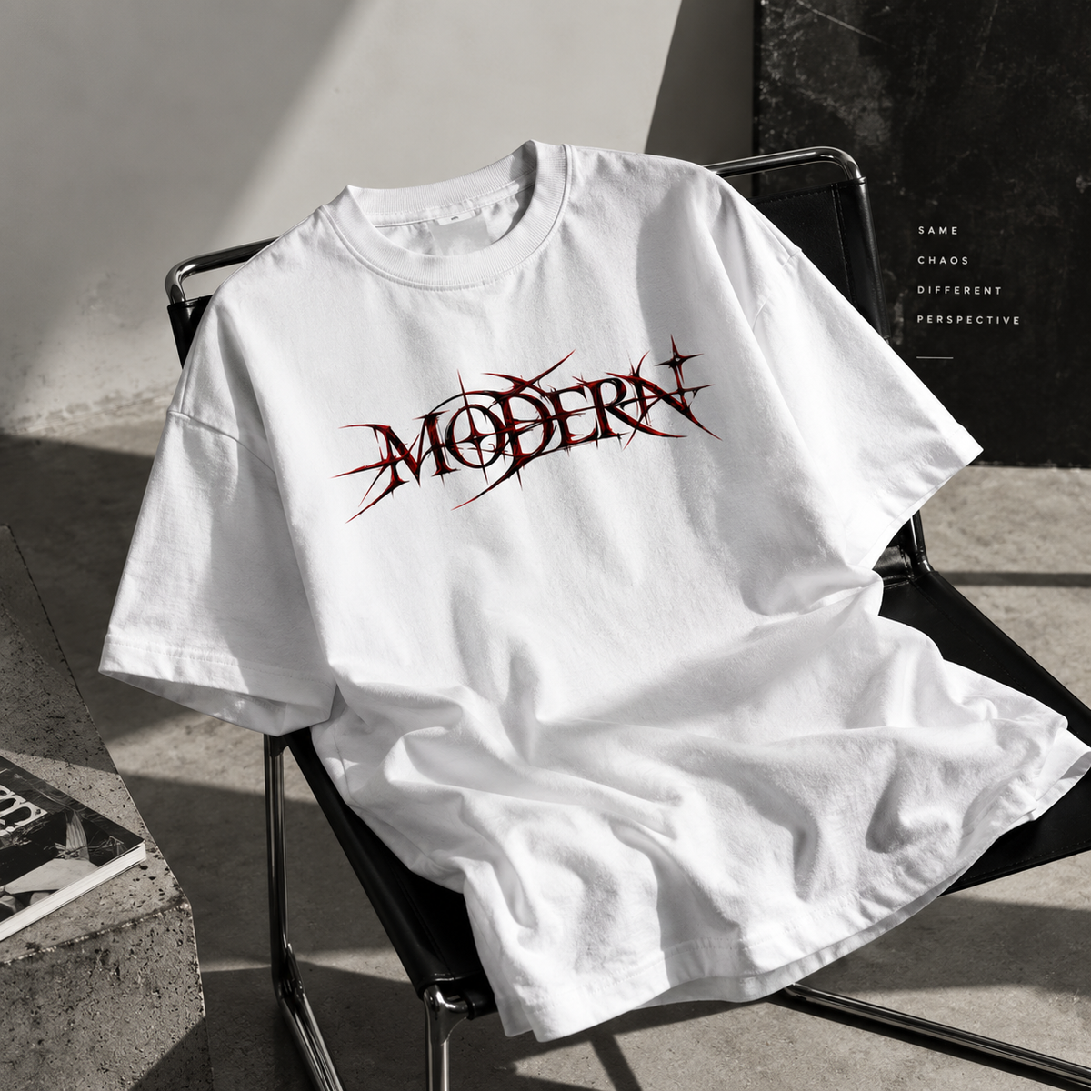 A white oversized T-shirt draped over a black metal chair in a sunlit studio setting. The shirt features a stylized dark red and black 'MODERN' graphic across the chest.
