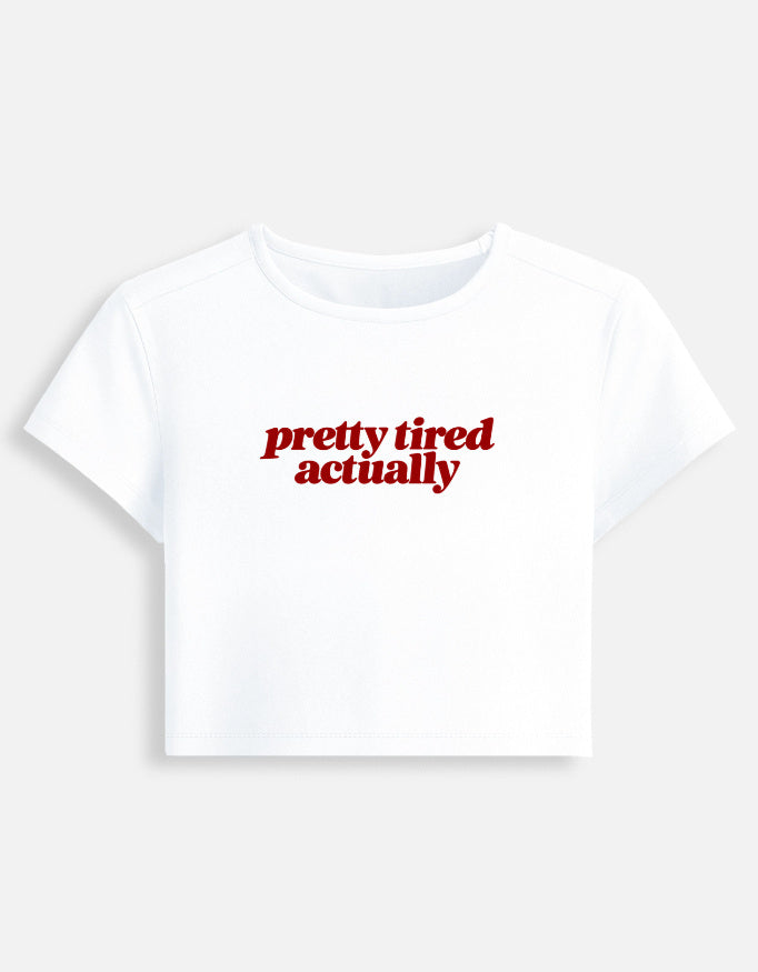 A white short-sleeve crop top t-shirt featuring the phrase 'pretty tired actually' printed in red serif text across the chest, displayed on a light gray background.