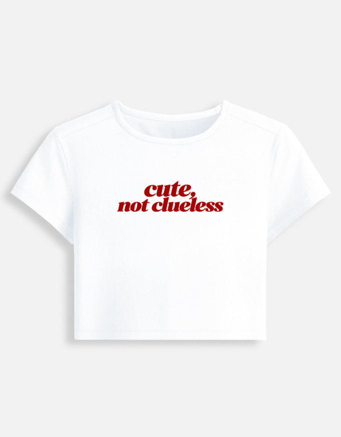 A white short-sleeve crop top t-shirt with the text 'cute, not clueless' printed in red across the chest, displayed on a light gray background.