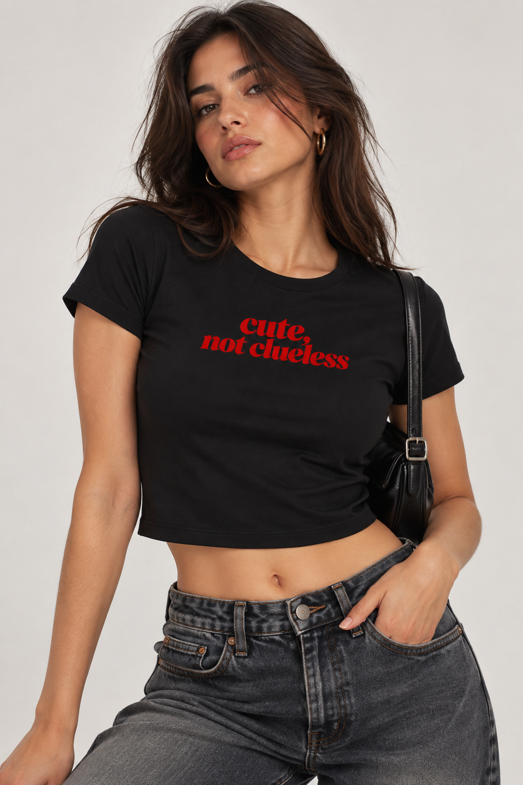 A woman wearing a black cropped short-sleeve t-shirt with the text 'cute, not clueless' printed in red across the chest, paired with dark gray jeans and a black shoulder bag, against a light gray studio background.