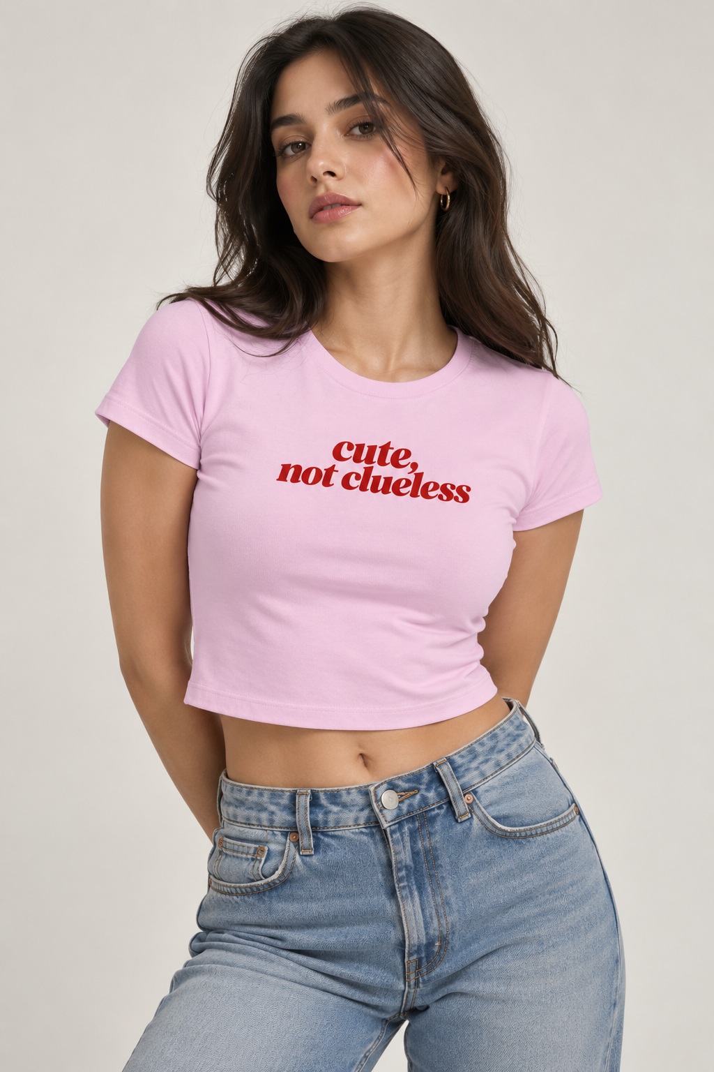 A woman wearing a light pink cropped t-shirt with the red text 'cute, not clueless' printed across the chest, paired with light blue jeans, set against a plain light gray background.