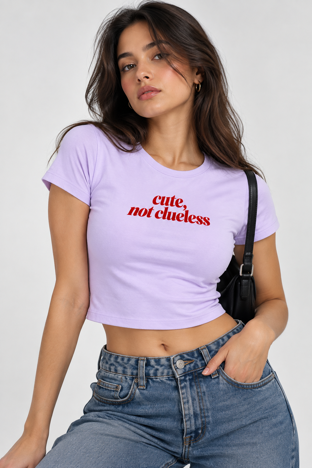 A woman wearing a light purple cropped t-shirt with the text 'cute, not clueless' printed in red across the chest, paired with blue jeans and a black shoulder bag, set against a plain light gray background.
