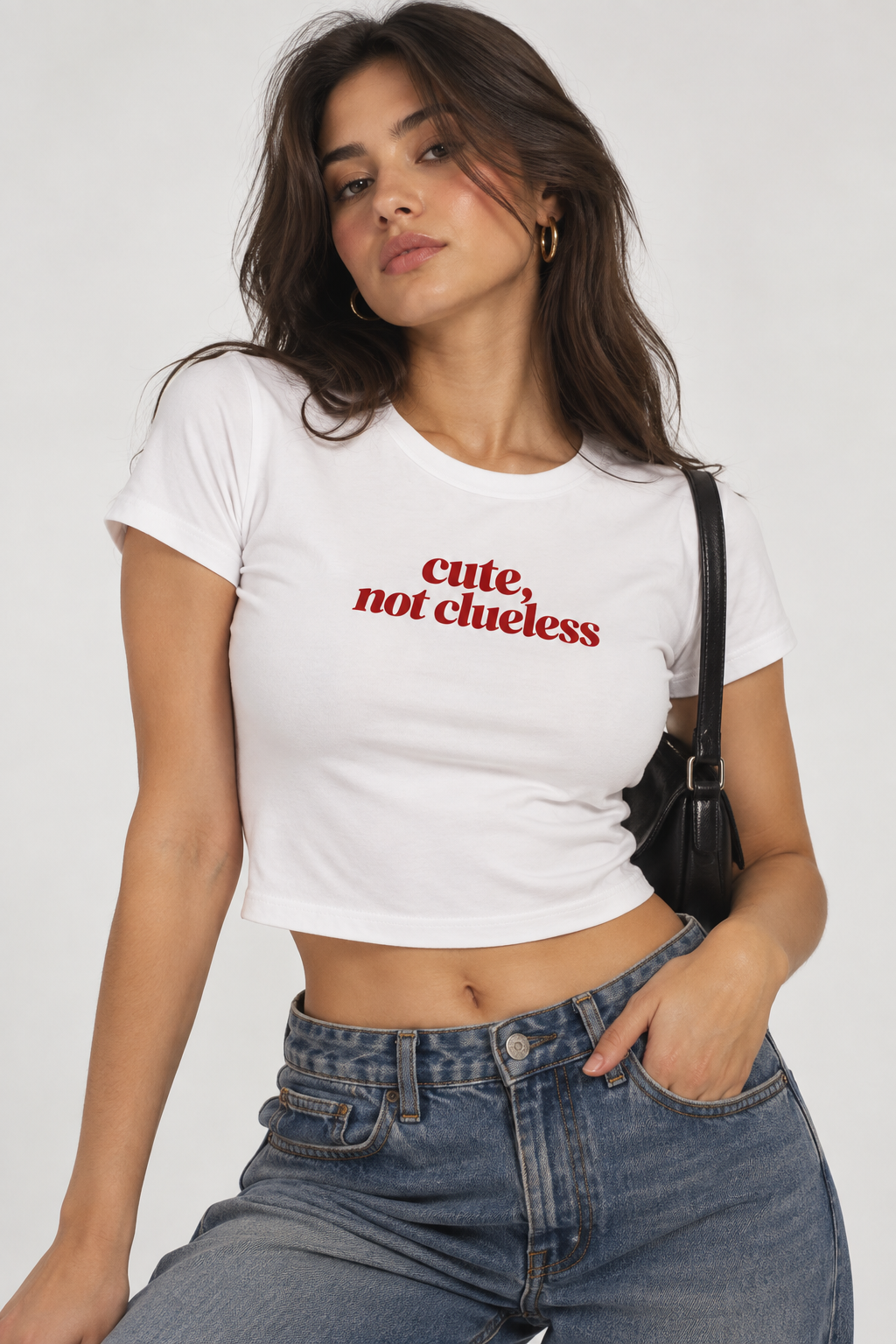 A woman wearing a white short-sleeve crop top with the red text 'cute, not clueless' printed on the chest, paired with blue jeans and a black shoulder bag, against a light gray studio background.