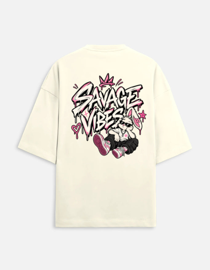 An oversized cream-colored t-shirt shown from the back, featuring a large graphic print with the text 'SAVAGE VIBES' in graffiti-style lettering, pink hearts, and a cartoon character wearing pink sneakers.