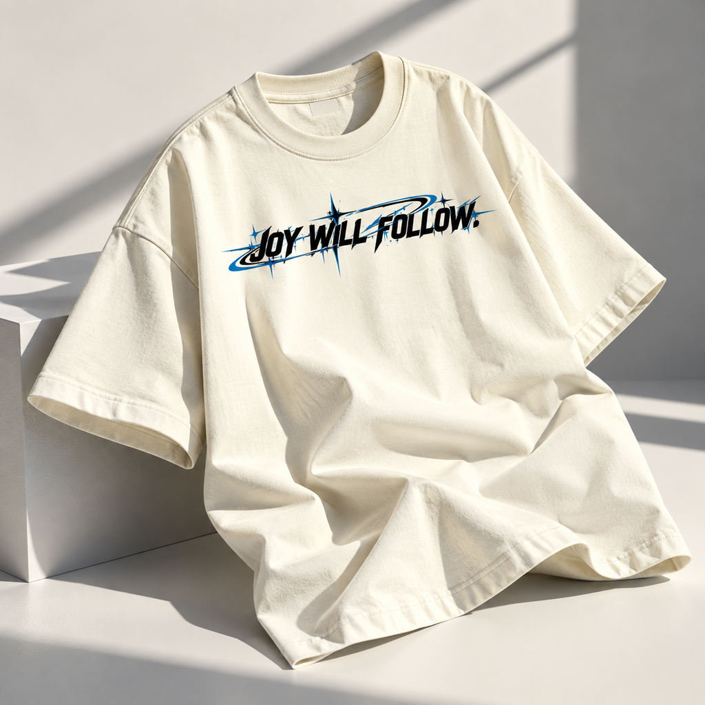 An oversized cream-colored t-shirt with a graphic print reading 'JOY WILL FOLLOW.' in black and blue, styled on a white geometric platform with warm natural shadows.