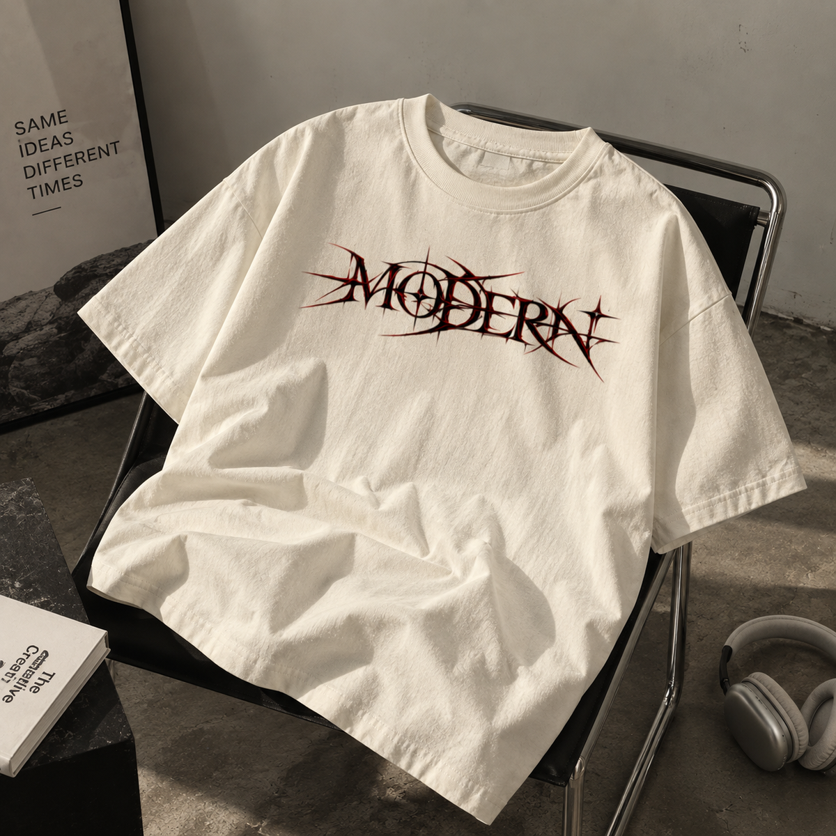 An oversized off-white t-shirt with a stylized black and red 'MODERN' graphic printed across the chest, styled on a black chair in a minimalist room setting with headphones nearby.