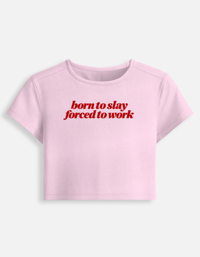 A light pink short-sleeve crop top t-shirt with the phrase 'born to slay forced to work' printed in red across the chest, displayed on a clean off-white background.
